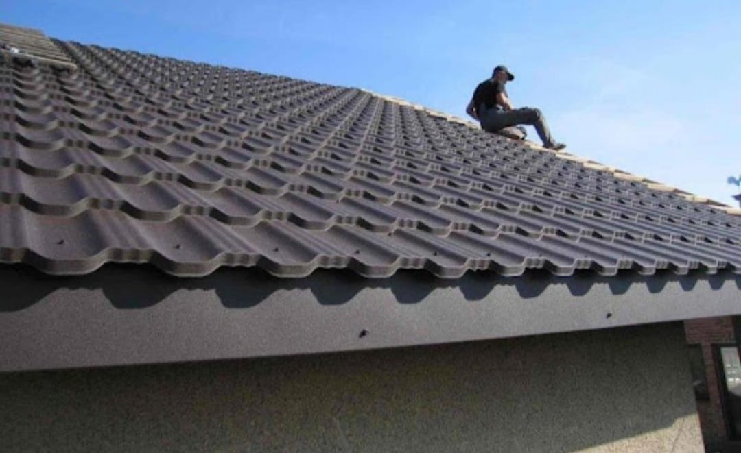 Pracownik siedzi na brązowym dachu z blachodachówki, widok z dołu na budynek z szarą elewacją i błękitne niebo.