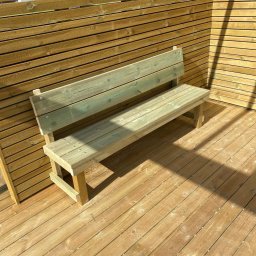 Bau mit uns ( Buduj z nami ) - Drewniana ławka z oparciem, wykonana z jasnego drewna, ustawiona na drewnianym tarasie, otoczona drewnianym ogrodzeniem z poziomych desek. Na podłodze widoczne ślady po obróbce drewna.