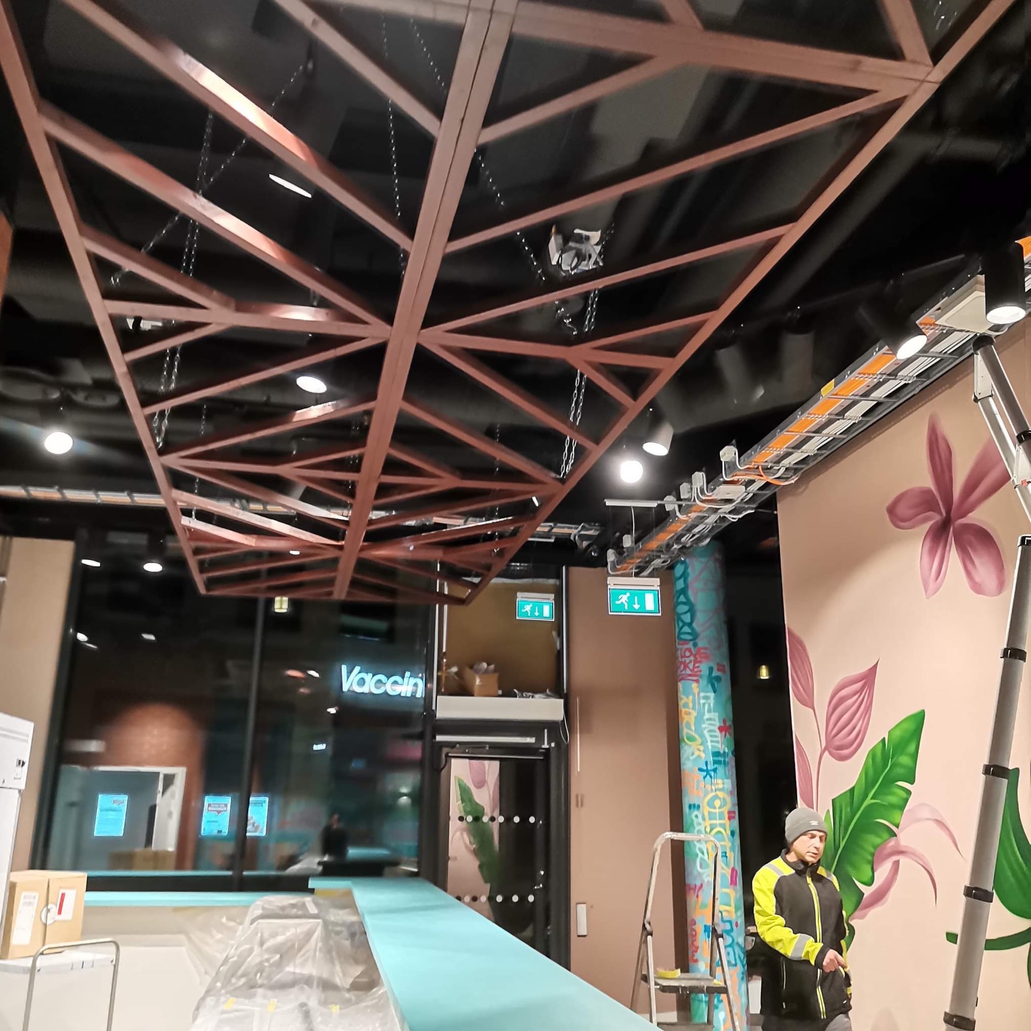 Montaż ażurowej konstrukcji sufitowej z drewna, widoczne przewody elektryczne, malowanie ścian z motywami roślinnymi, pracownik w odblaskowej kurtce i czapce, zabezpieczona podłoga.