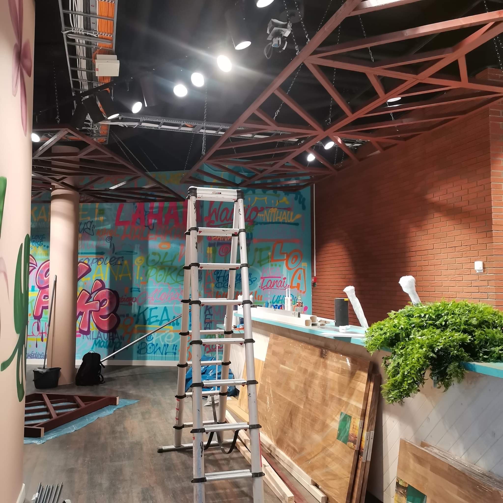 Wnętrze lokalu w trakcie prac wykończeniowych: ściana z kolorowym graffiti, ceglana ściana, drewniana konstrukcja sufitu, drabina rozstawiona na podłodze, materiały budowlane i rośliny dekoracyjne.