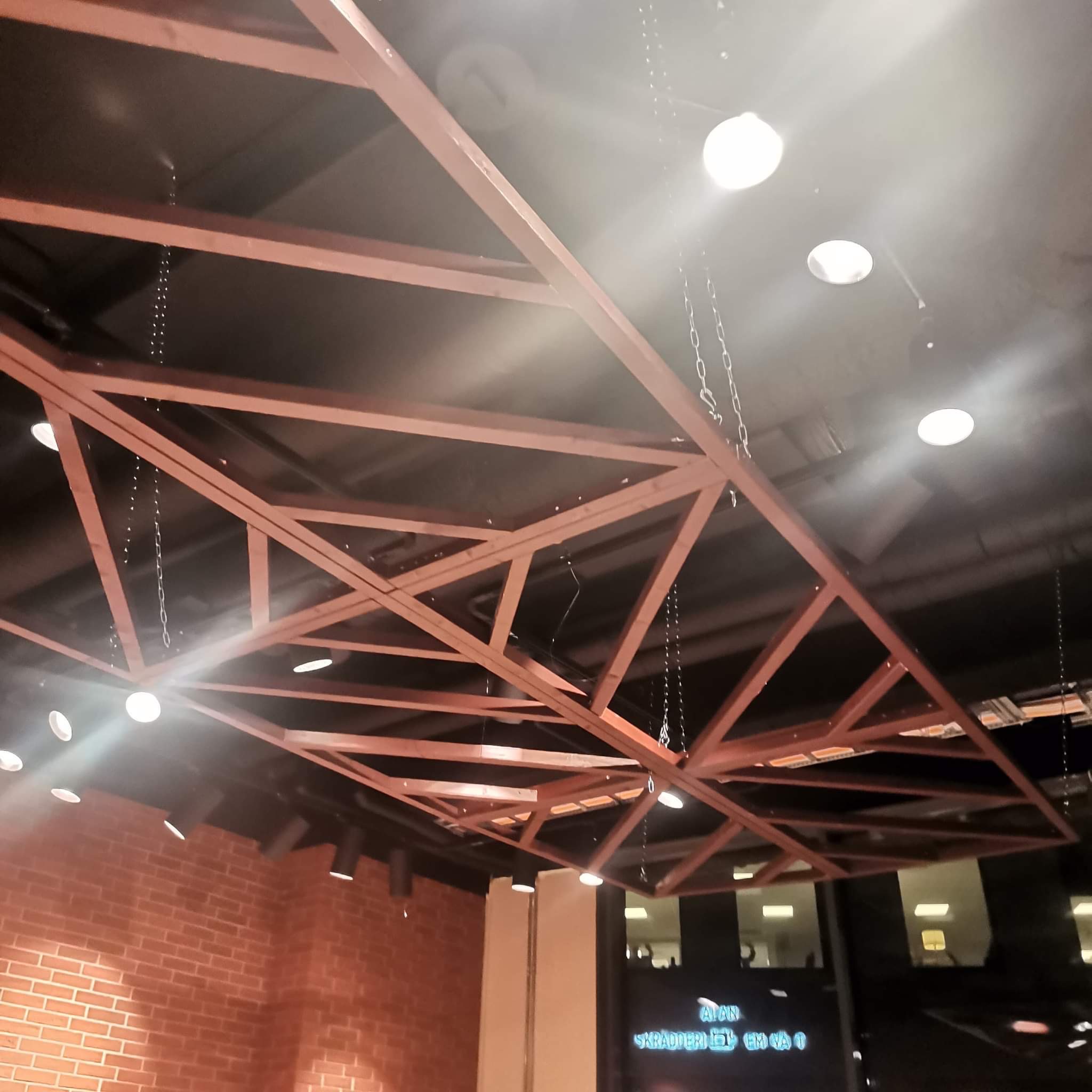 Drewniana konstrukcja sufitowa z oświetleniem punktowym i łańcuchami, widok od dołu, ceglana ściana w tle.