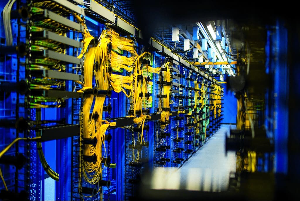 Rząd serwerów w serwerowni z niebieskimi szafami i żółtymi kablami światłowodowymi, widok z perspektywy korytarza.