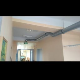 Brak - Szare, spiralne rury wentylacyjne zamontowane pod sufitem w korytarzu budynku z beżowymi ścianami i zielonym znakiem ewakuacyjnym nad drzwiami.