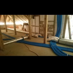 Brak - Instalacja przewodów wentylacyjnych i niebieskich rur w surowym wnętrzu poddasza z drewnianą konstrukcją, widoczne fragmenty ocieplonych ścian.