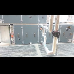 Brak - Szara centrala wentylacyjna VTS z rurami i pompą obiegową Grundfos, umieszczona na metalowej kratownicy podłogowej.