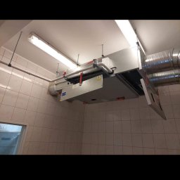 Brak - Centrala wentylacyjna podwieszona pod sufitem w pomieszczeniu wyłożonym kafelkami, widoczne rury i przewody wentylacyjne.