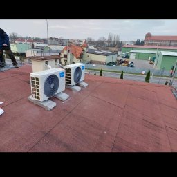 Brak - Dwie jednostki zewnętrzne klimatyzacji Midea zamontowane na płaskim dachu pokrytym czerwoną papą, widok na zabudowę miejską w tle.