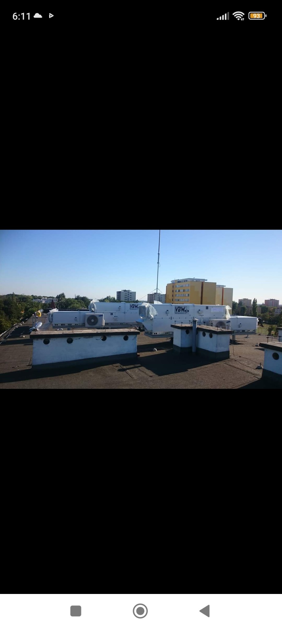 Instalacja klimatyzacji na dachu budynku, widoczne jednostki zewnętrzne klimatyzatorów z logo VBW, częściowo owinięte folią ochronną, na tle zabudowy miejskiej.