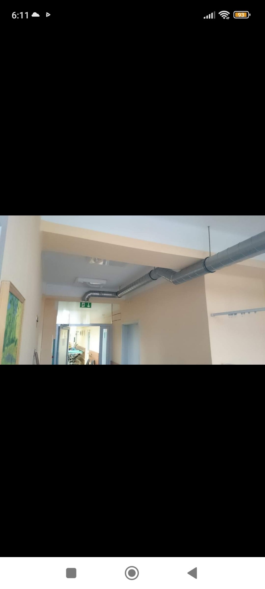 Szare, spiralne rury wentylacyjne zamontowane pod sufitem w korytarzu budynku z beżowymi ścianami i zielonym znakiem ewakuacyjnym nad drzwiami.