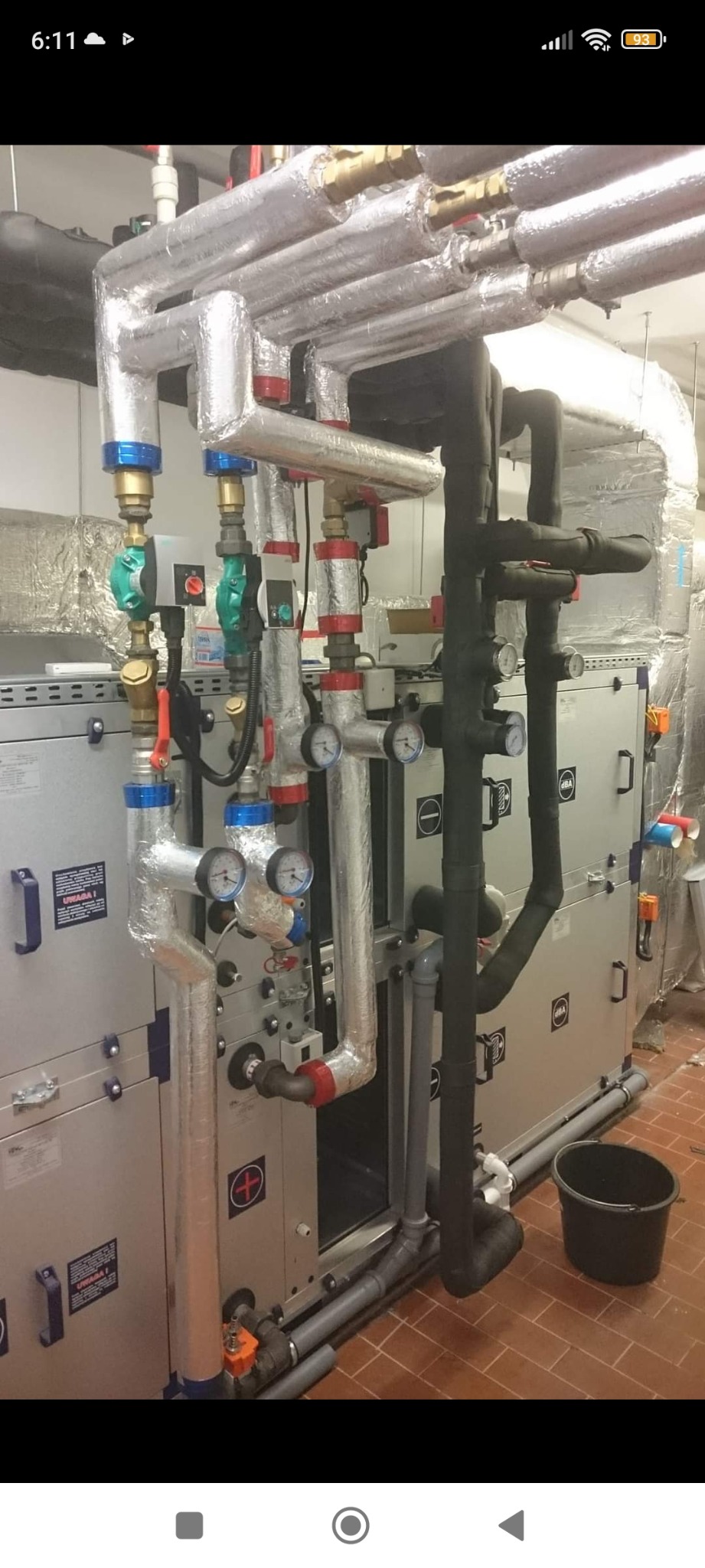 Złożona instalacja rur i urządzeń wentylacyjnych w pomieszczeniu technicznym, z rurami izolowanymi folią aluminiową i czarną pianką, widoczne manometry i zawory, szare obudowy urządzeń z naklejkami...