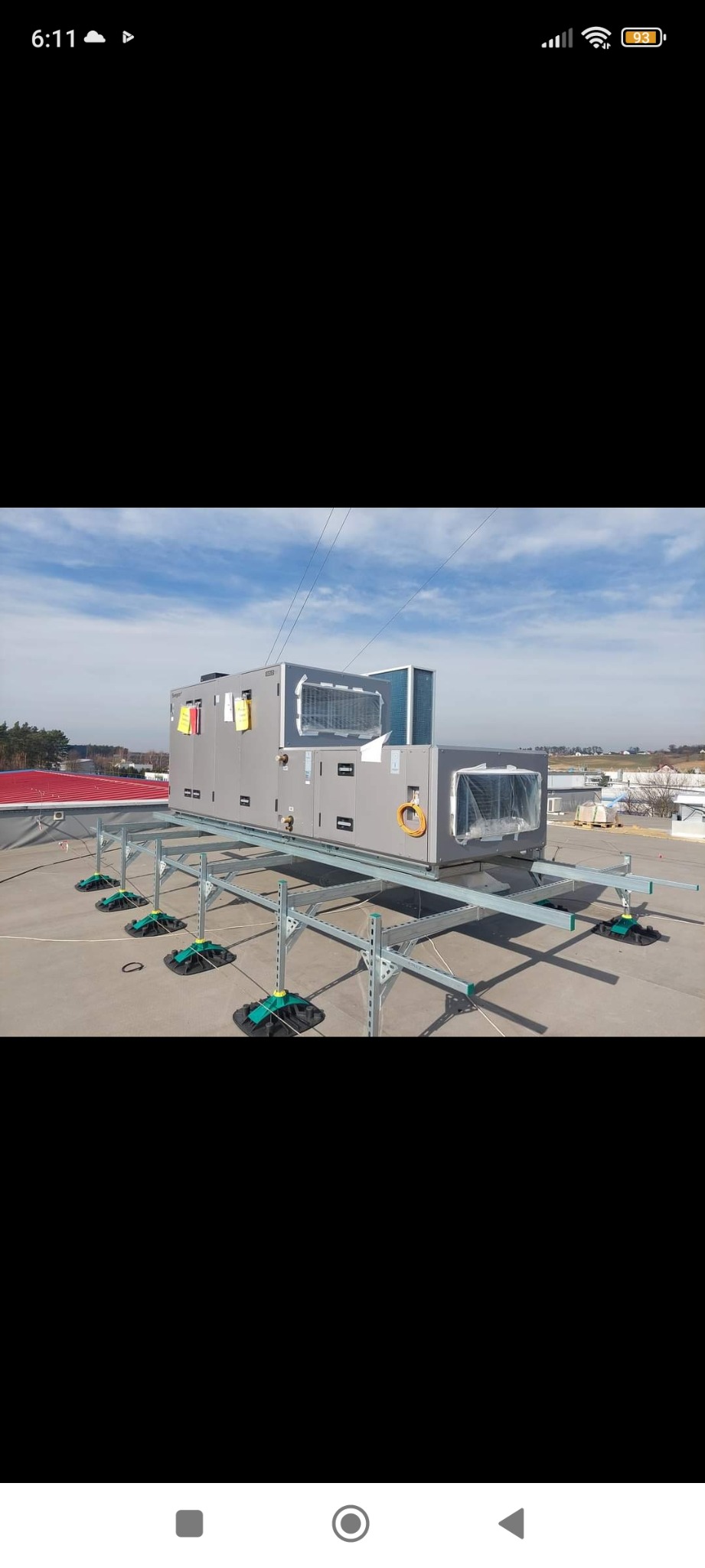 Szare jednostki klimatyzacyjne na dachu budynku, zamontowane na metalowej konstrukcji z zielonymi podkładkami antywibracyjnymi, widok z boku, w tle błękitne niebo i zabudowania.