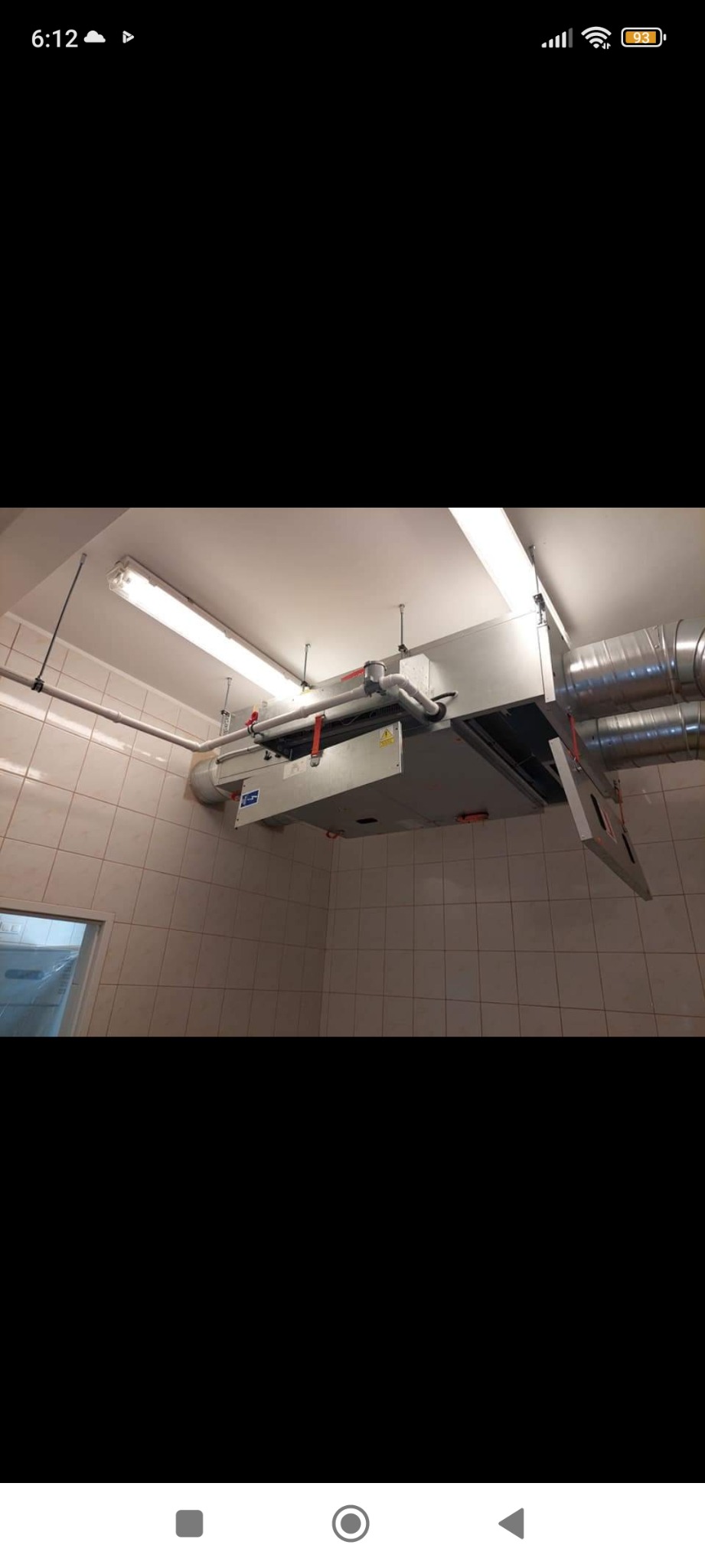 Centrala wentylacyjna podwieszona pod sufitem w pomieszczeniu wyłożonym kafelkami, widoczne rury i przewody wentylacyjne.