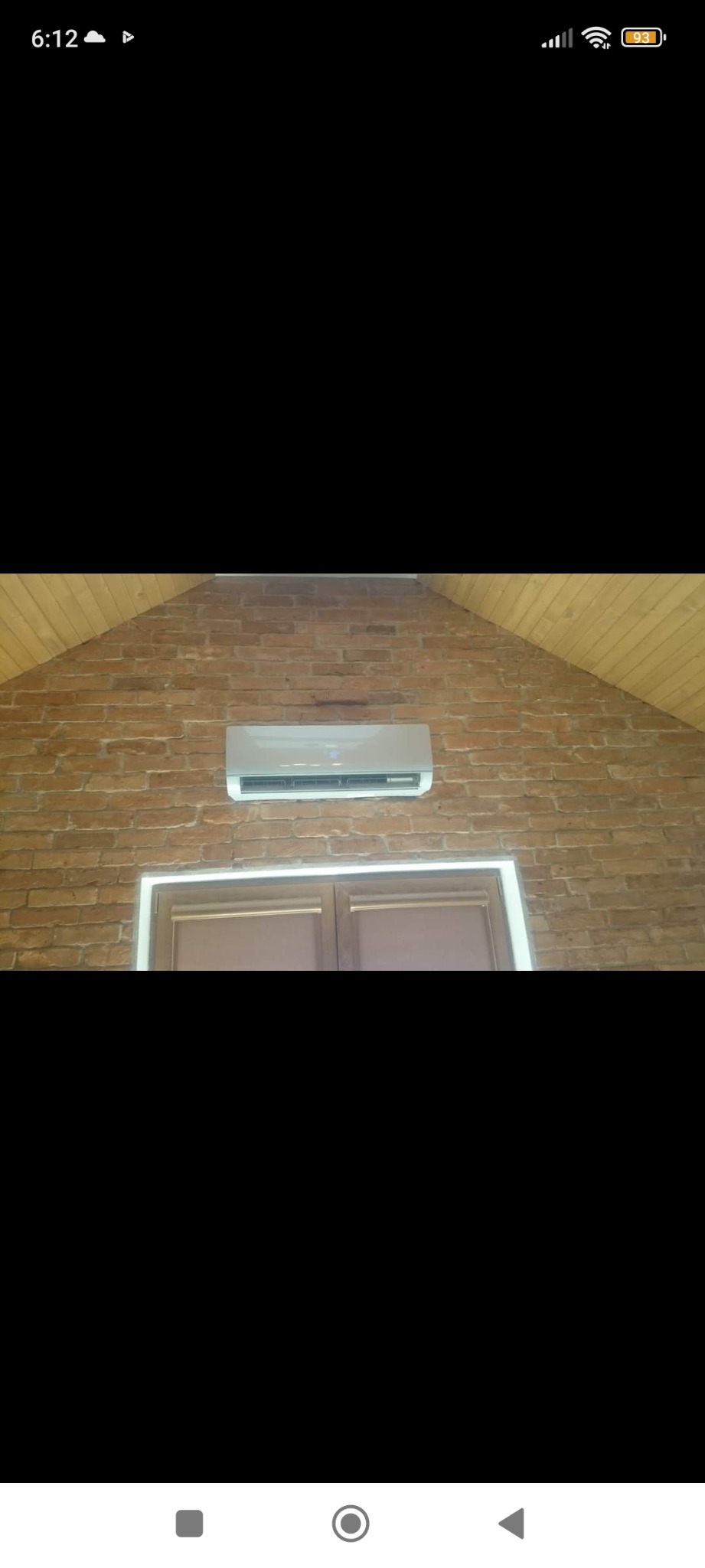 Klimatyzator ścienny zamontowany na ceglanej ścianie szczytowej, widoczny nad drewnianymi drzwiami z ościeżnicą, pod drewnianą boazerią dachową.