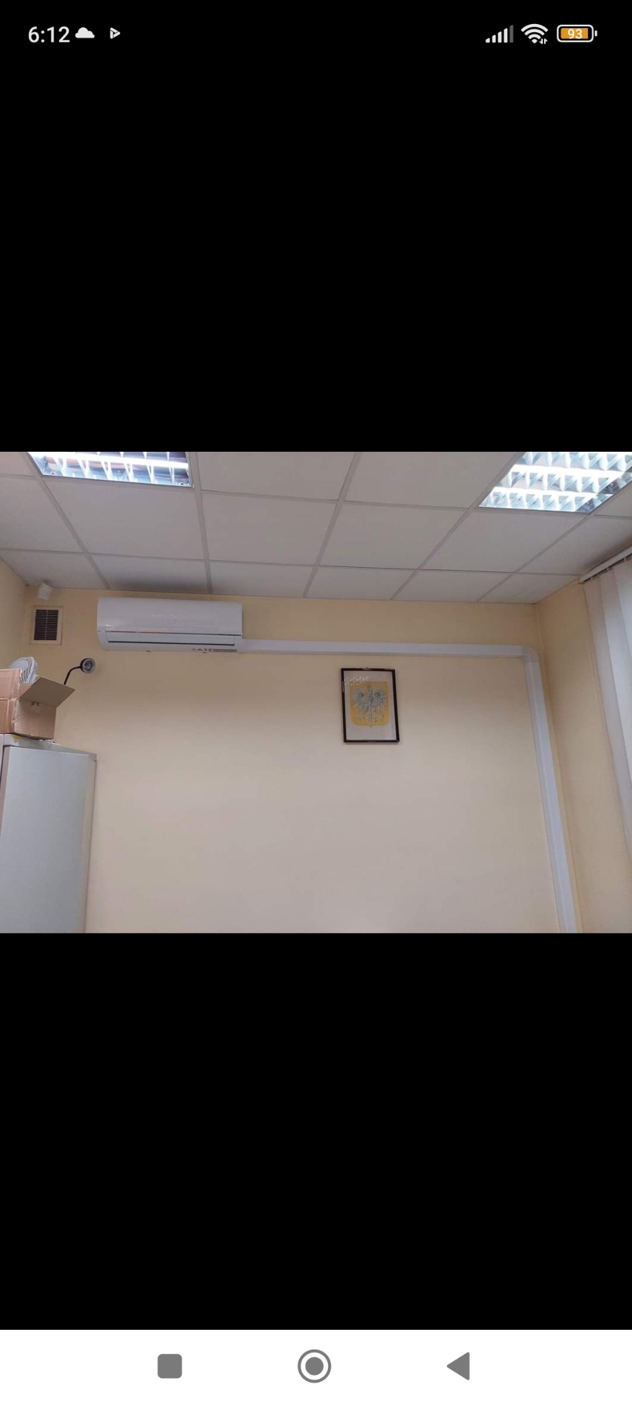 Biała klimatyzacja ścienna w biurze, widok z dołu, obok szafka z kartonem i herb w ramce na ścianie.