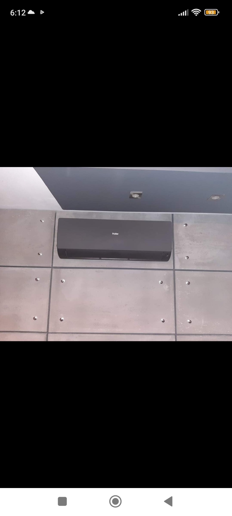 Czarny klimatyzator Haier zamontowany na ścianie z betonowych płyt z metalowymi mocowaniami, widok z dołu, oświetlenie sufitowe.