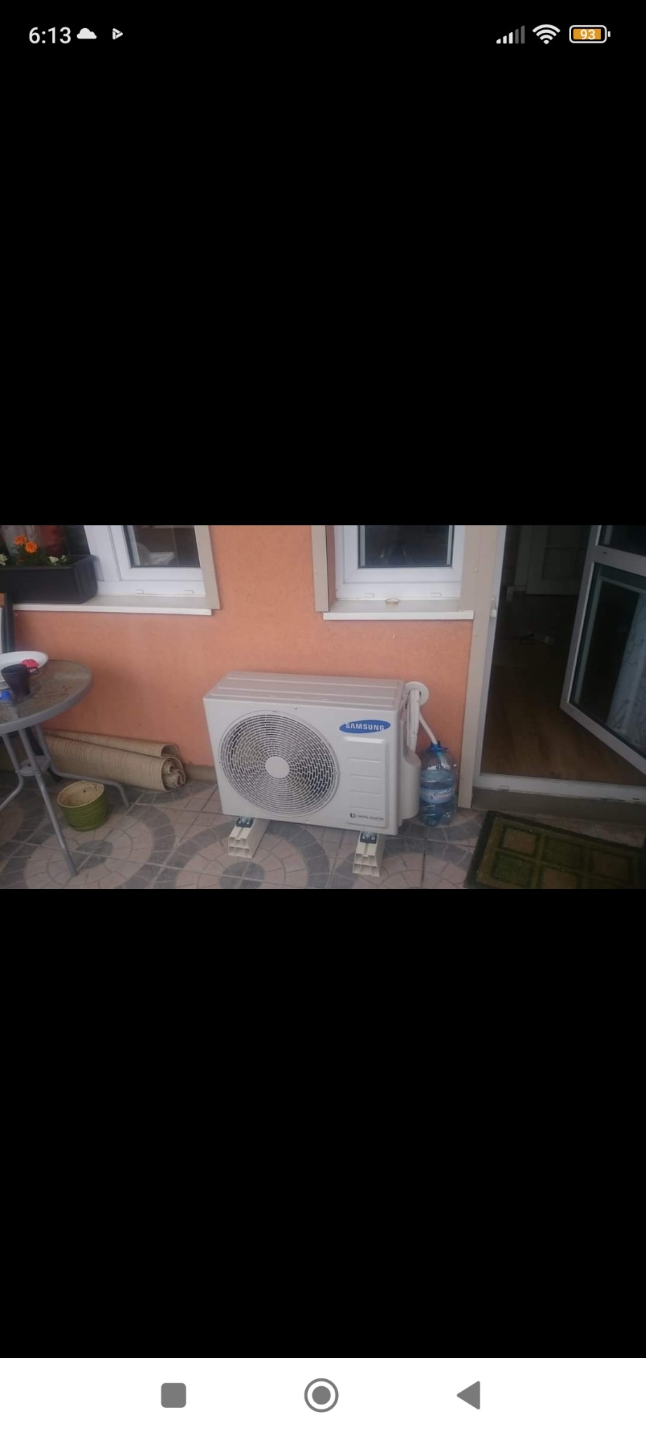 Zewnętrzna jednostka klimatyzacji Samsung Digital Inverter zamontowana na plastikowych wspornikach przy pomarańczowej ścianie budynku, z butelką wody obok.