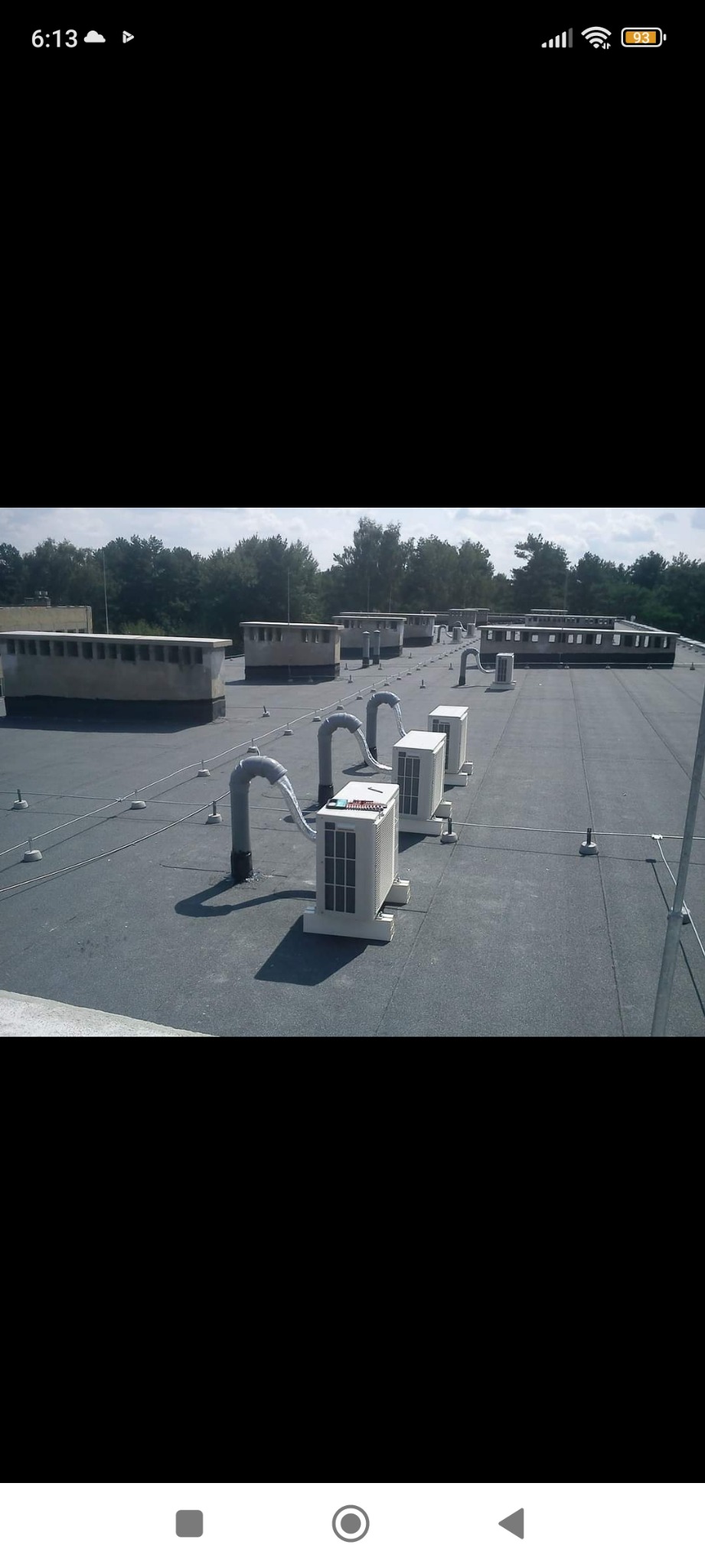 Rząd jednostek klimatyzacyjnych na płaskim dachu budynku, z widocznymi przewodami wentylacyjnymi i kominami w tle.