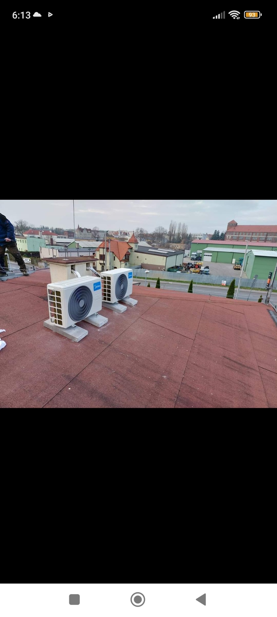 Dwie jednostki zewnętrzne klimatyzacji Midea zamontowane na płaskim dachu pokrytym czerwoną papą, widok na zabudowę miejską w tle.