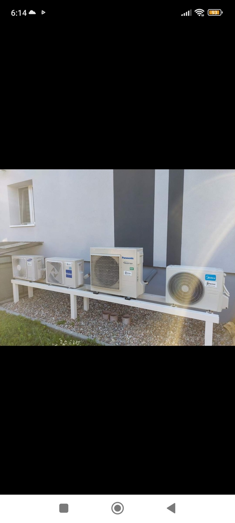 Zewnętrzne jednostki klimatyzacyjne różnych marek (Samsung, Haier, Panasonic, Midea) ustawione na białej metalowej konstrukcji przed budynkiem z szaro-białymi pasami, widoczne także kamienie...