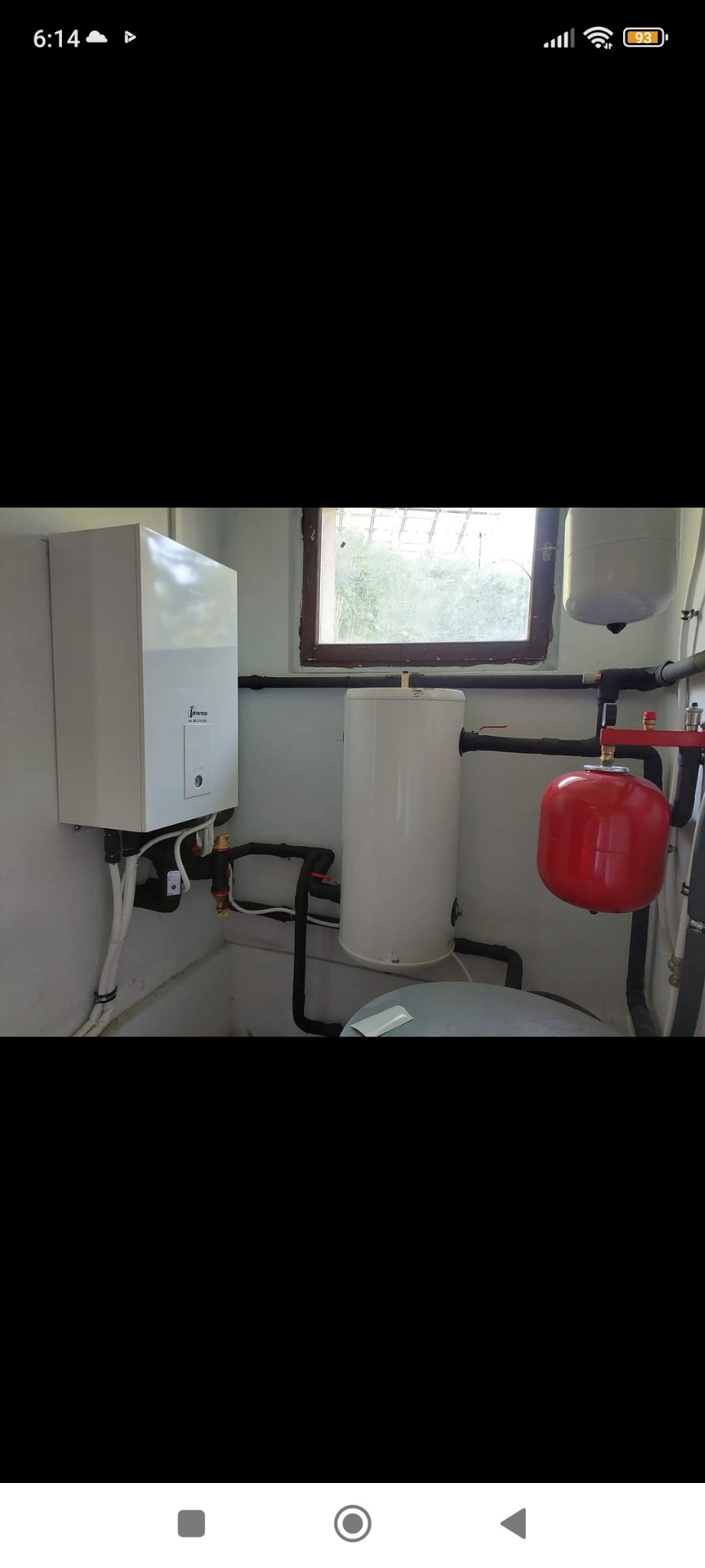 Biała pompa ciepła wisząca na ścianie w kotłowni, obok pionowy zbiornik akumulacyjny i czerwony zbiornik wyrównawczy, widoczne rury instalacji grzewczej i małe okno.