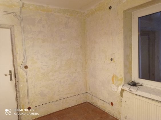 Ściana w trakcie remontu z poprowadzonymi przewodami elektrycznymi w listwach przypodłogowych i puszkami instalacyjnymi, widoczne ślady po starych tapetach i gniazdkach.