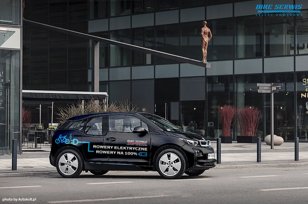 Czarny samochód elektryczny z naklejką reklamową Bike Serwis na tle nowoczesnego budynku biurowego z rzeźbą