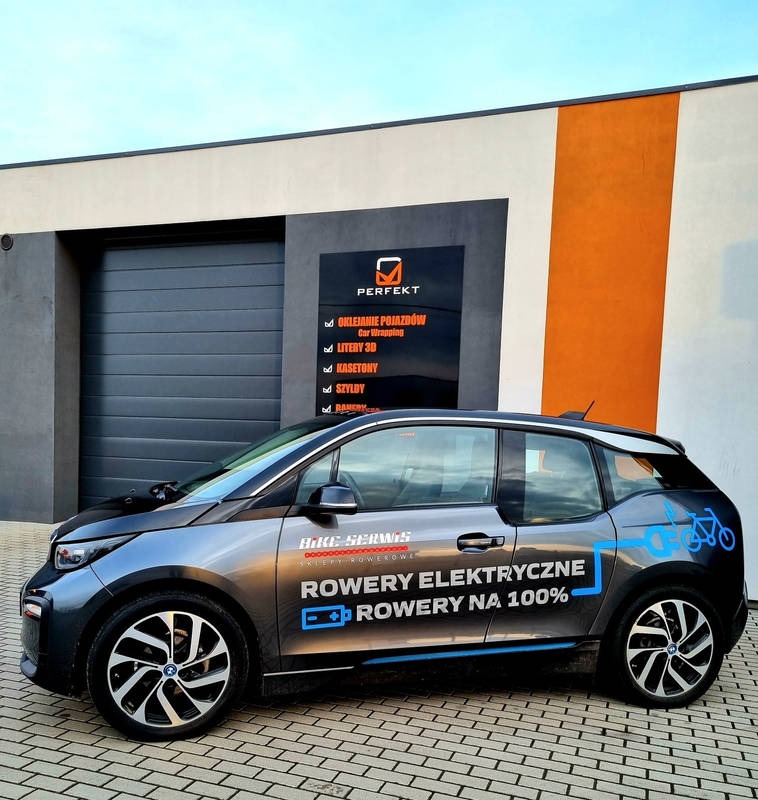 Naklejka reklamowa Bike Serwis z rowerem elektrycznym na samochodzie BMW i3, wykonana przez firmę Perfekt, widoczna na tle szaro-pomarańczowej ściany budynku z zamkniętą bramą garażową.