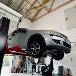 Srebrny samochód osobowy Opel podniesiony na dwukolumnowym podnośniku w warsztacie samochodowym z widocznymi narzędziami i wyposażeniem, w tle białe ściany i brama garażowa.
