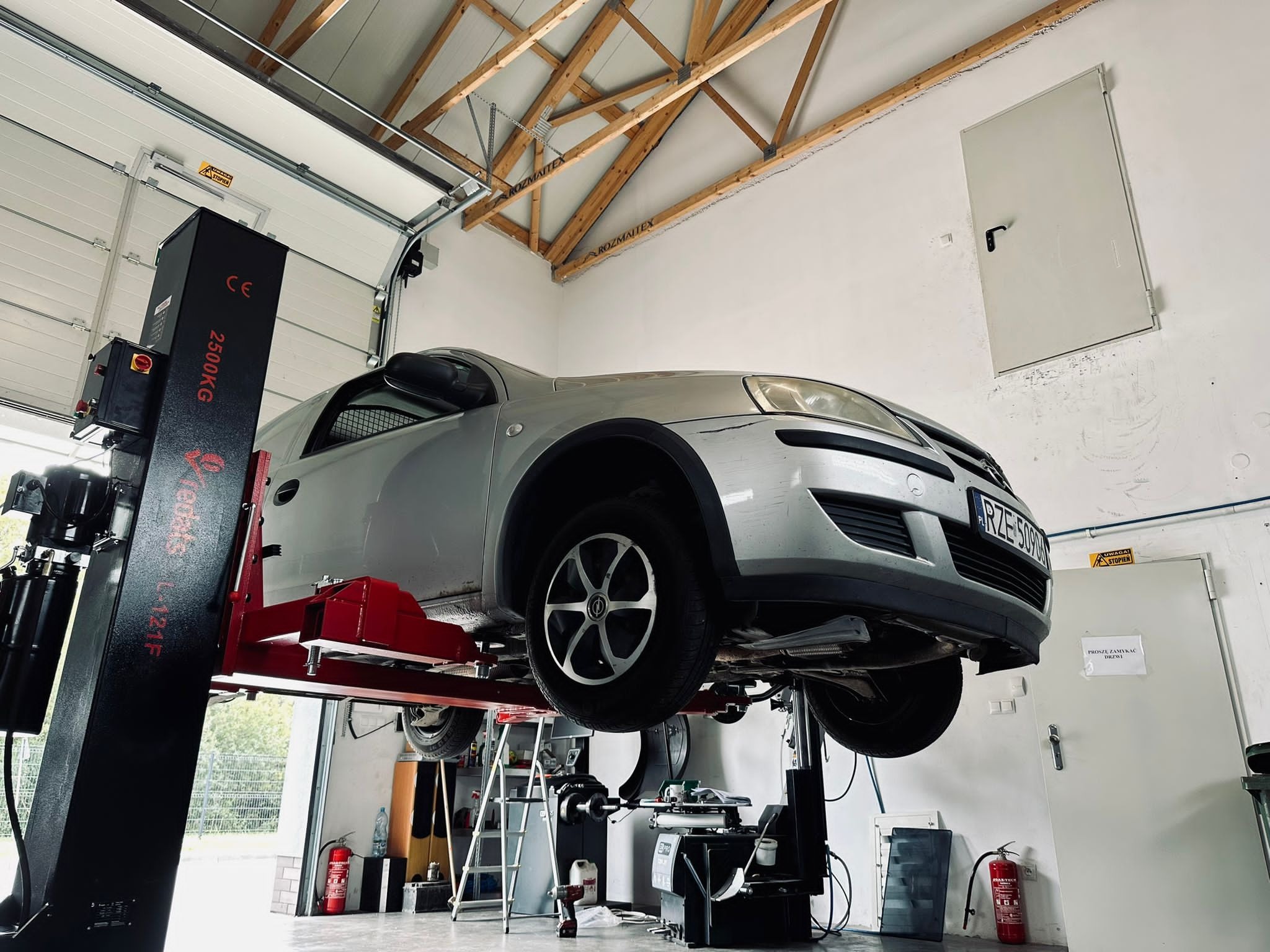 Srebrny samochód osobowy Opel podniesiony na dwukolumnowym podnośniku w warsztacie samochodowym z widocznymi narzędziami i wyposażeniem, w tle białe ściany i brama garażowa.