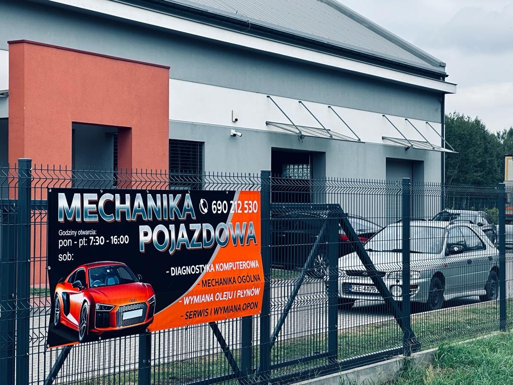 Baner reklamowy warsztatu samochodowego z czerwonym sportowym autem i informacjami o usługach, zawieszony na ogrodzeniu, z budynkiem warsztatu i zaparkowanymi samochodami w tle.