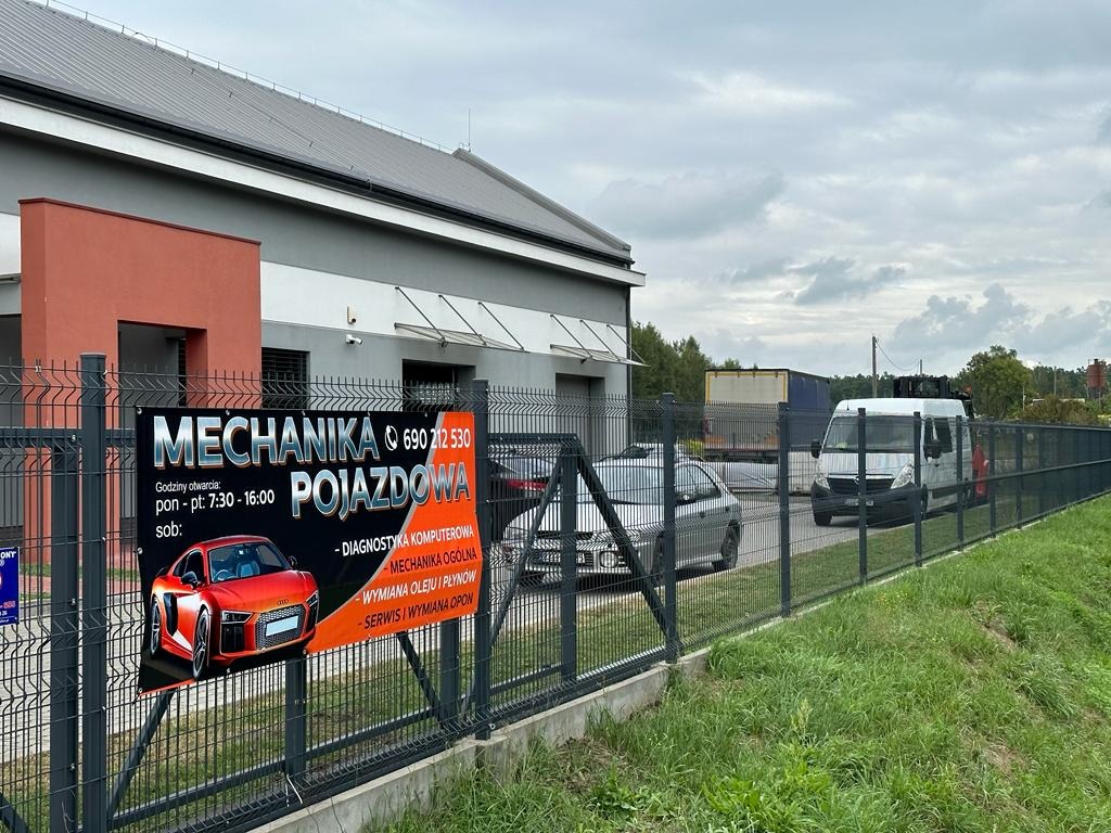 Baner reklamowy warsztatu samochodowego z godzinami otwarcia i zakresem usług, umieszczony na metalowym ogrodzeniu, z widocznymi pojazdami w tle