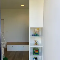 Biała szafa z półkami w pokoju dziecięcym, widoczne zabawki na półkach, drewniana podłoga, w tle huśtawka hamak.