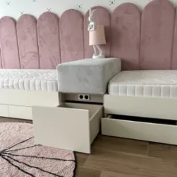 Pokój dziecięcy z dwoma łóżkami z szufladami, tapicerowanym zagłówkiem w pudrowym różu, pufą i lampką nocną w kształcie królika, na tle ściany z motywem króliczych buziek.