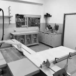 Warsztat stolarski w czerni i bieli: piła stołowa tnie płytę z jasnego drewna, odciąg wiórów, narzędzia na ścianie, stół roboczy z trzema krzesłami z wikliny w środku, szafka na kółkach z szufladami.