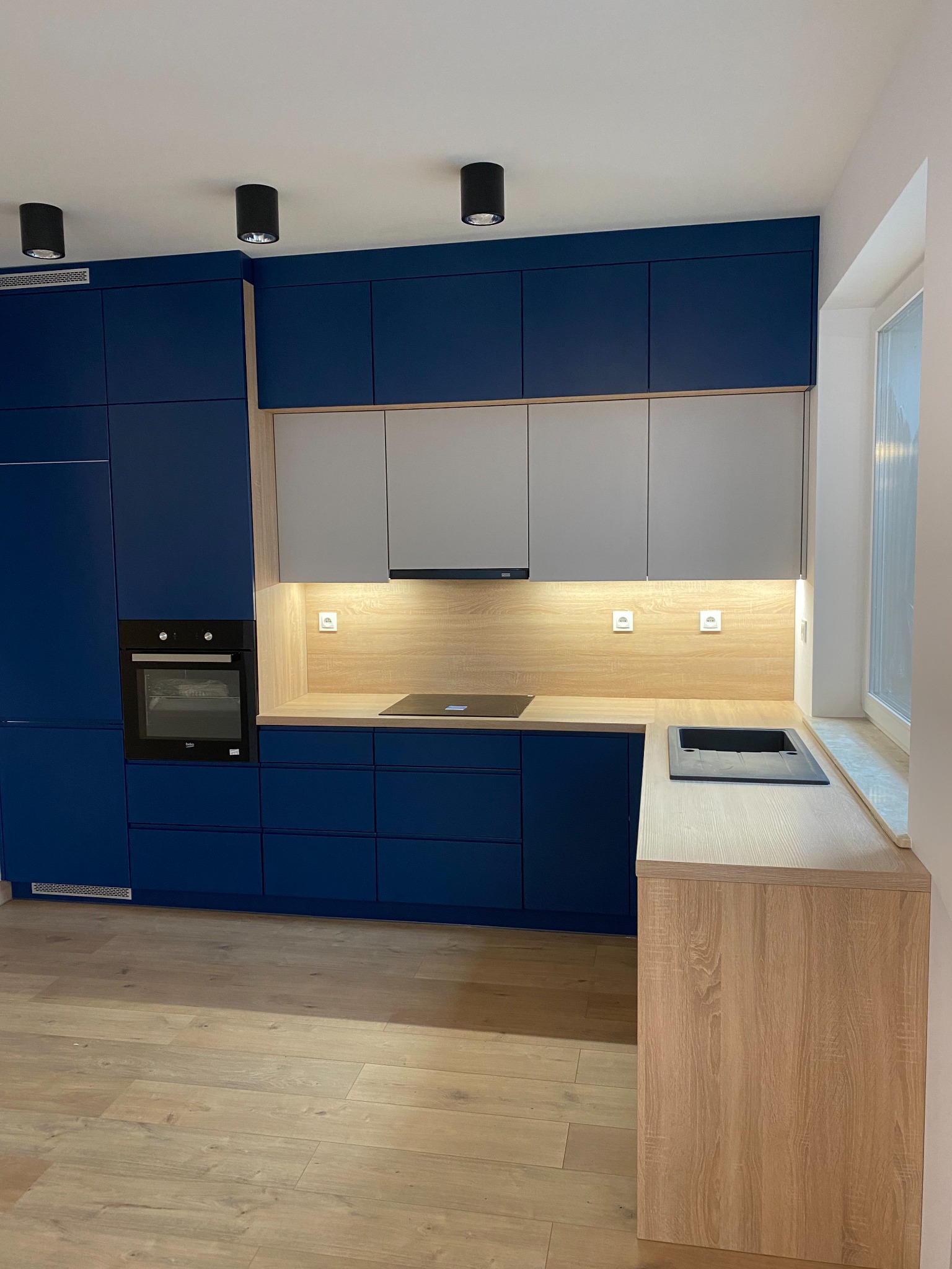 Nowoczesna kuchnia z granatowymi szafkami dolnymi i szarymi górnymi, drewniany blat roboczy z wbudowaną płytą indukcyjną i zlewozmywakiem, oświetlenie podszafkowe, minimalistyczne czarne lampy...