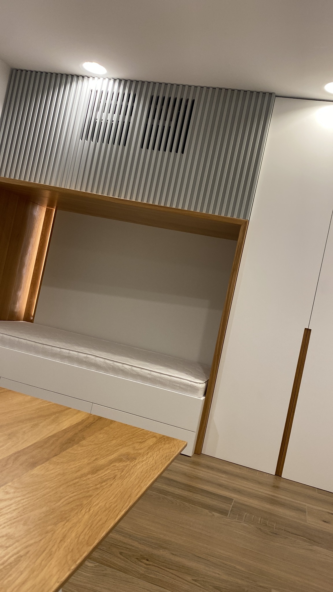 Biała szafa wnękowa z drewnianą wnęką i oświetleniem LED, nad nią szara dekoracyjna ściana z pionowych listew, fragment blatu drewnianego stołu na pierwszym planie.