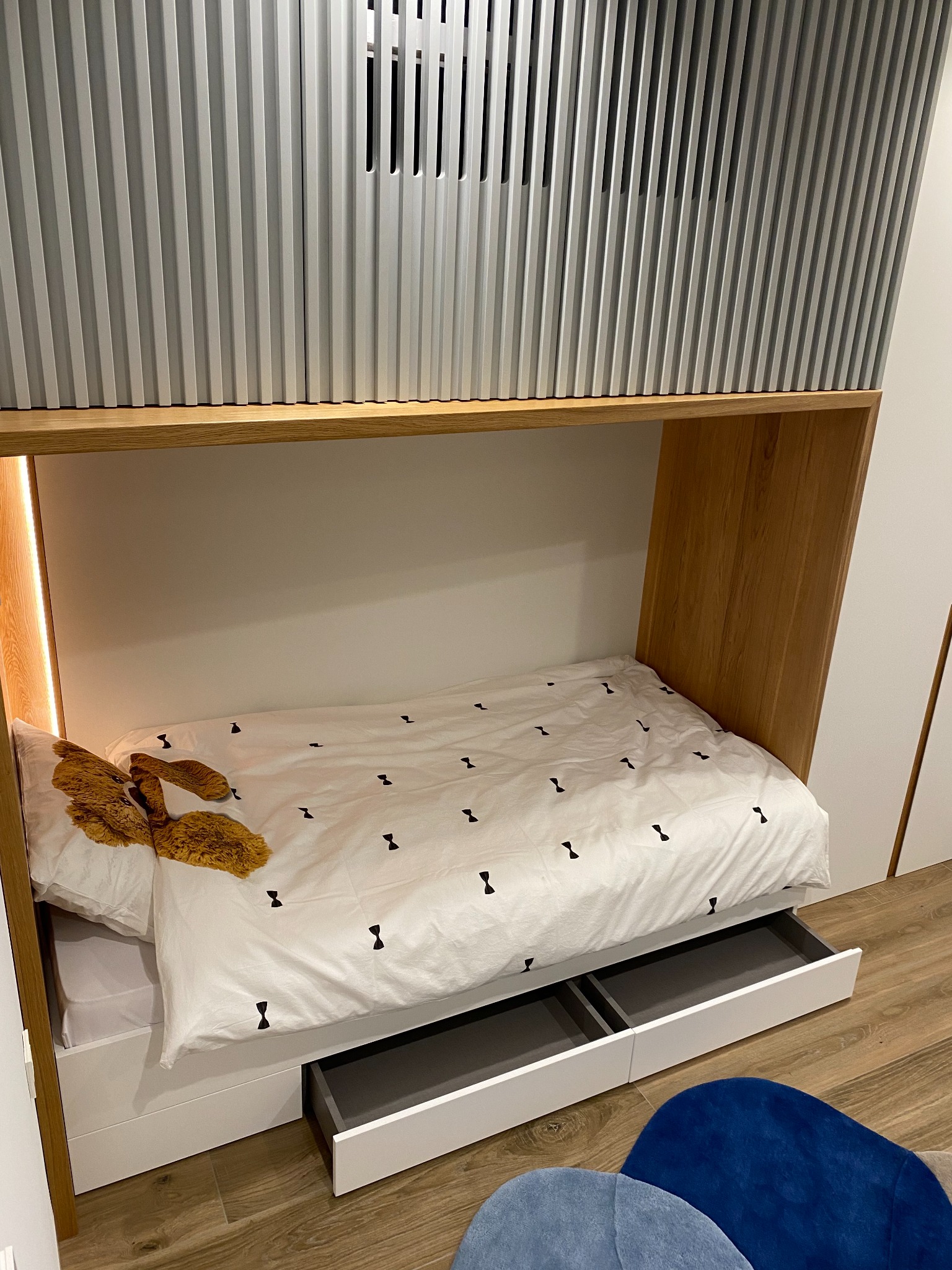 Wnęka z łóżkiem i wysuwanymi szufladami, wykończona drewnem, z białym zagłówkiem z pionowymi panelami i pościelą w czarne muszki. Na łóżku pluszowy miś.