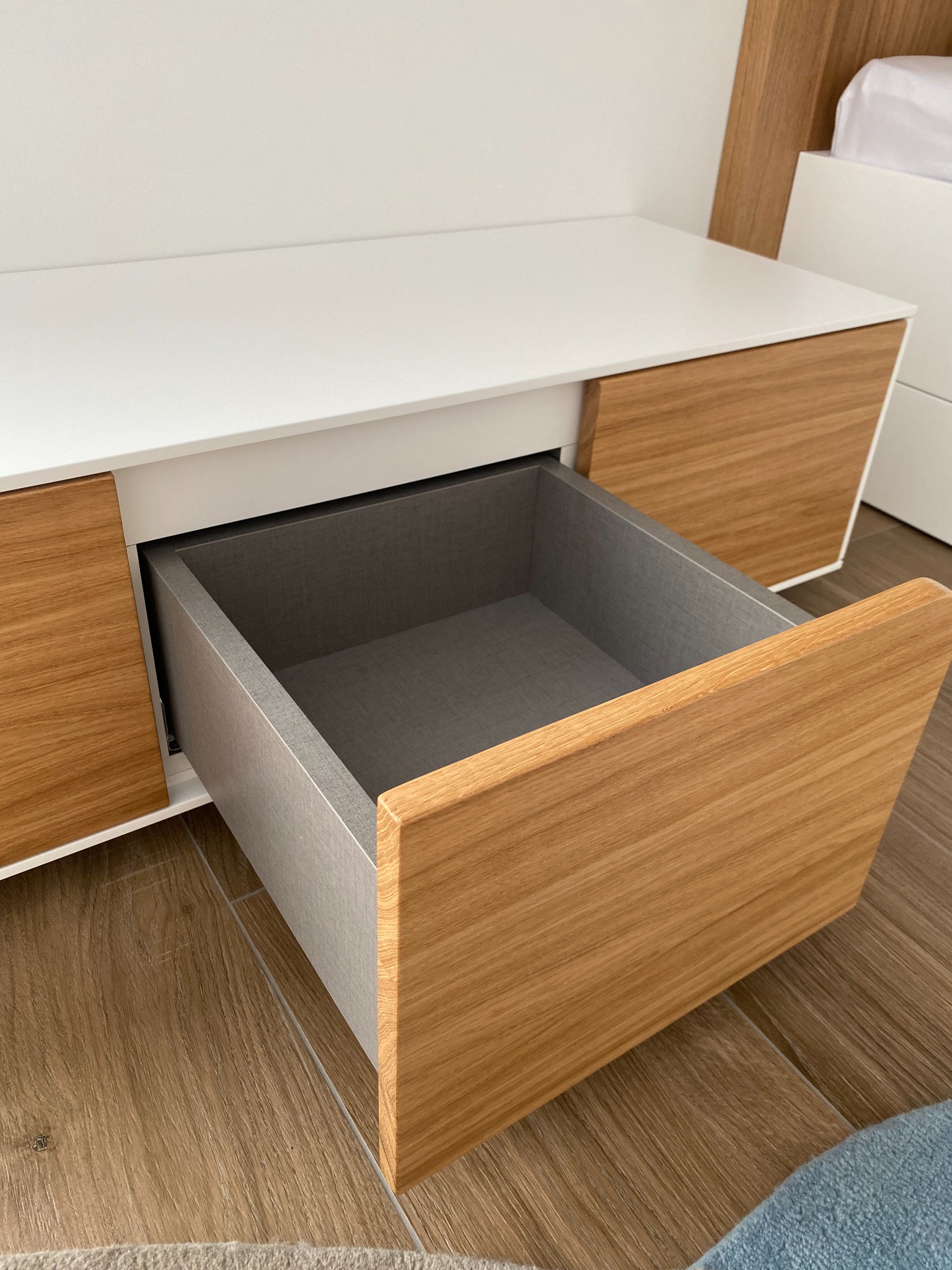 Wysuwana szuflada z drewnianym frontem i szarym wnętrzem, część mebla z białym blatem, widoczna podłoga z paneli.