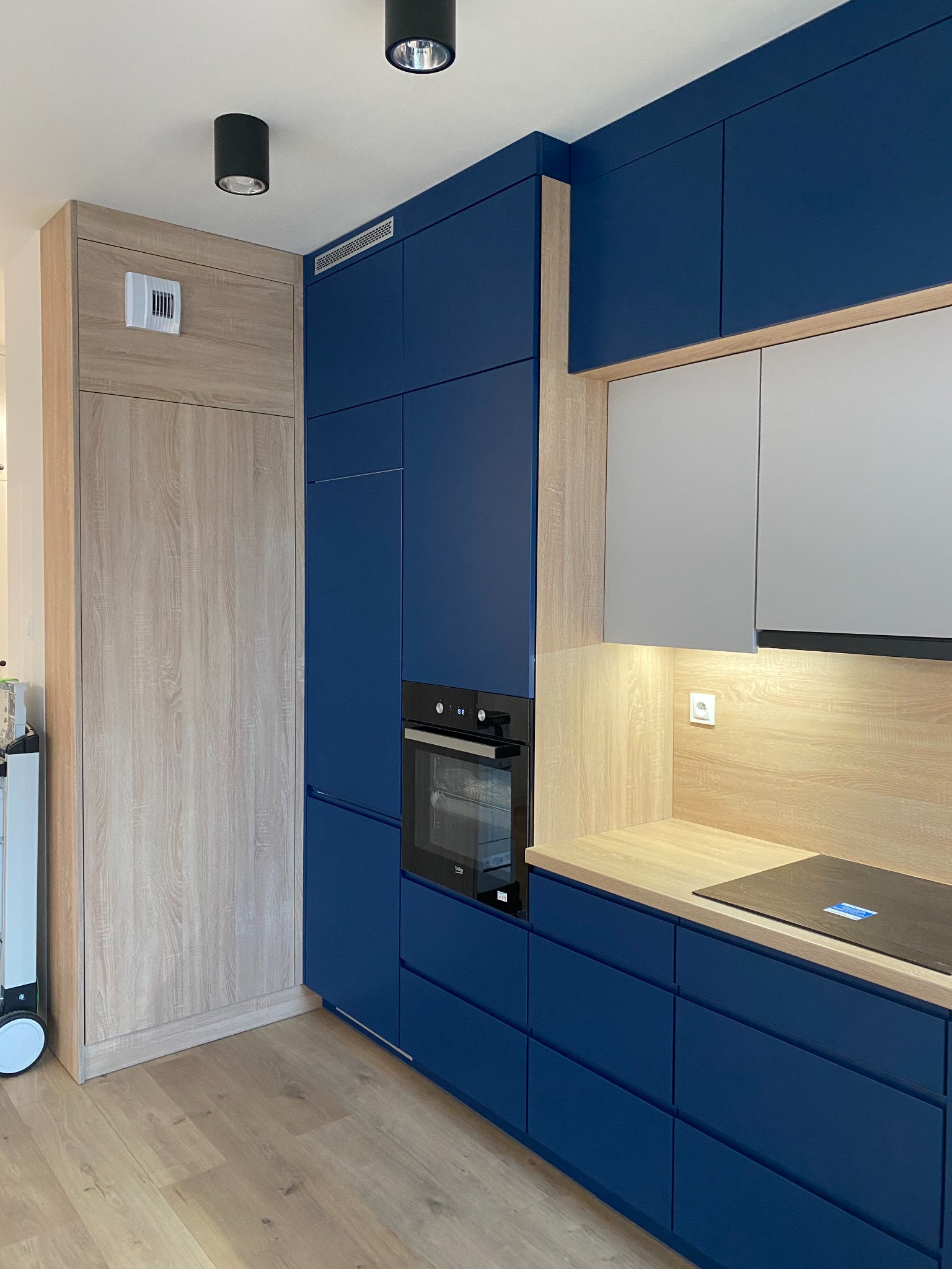 Nowoczesna kuchnia z granatowymi frontami szafek, drewnianym blatem i jasnoszarymi szafkami wiszącymi, oświetlona punktowymi lampami sufitowymi, z widocznym piekarnikiem i płytą indukcyjną.