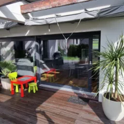 Zadaszony taras z przesuwnymi drzwiami, osłonięty szarą roletą zwijaną i zasłoną, z widocznymi roślinami w donicach i dziecięcym stolikiem z krzesełkami.