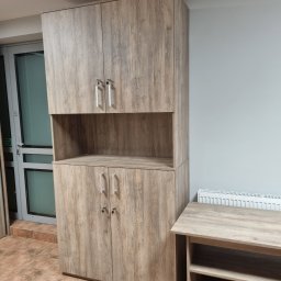 MEBLOTER Paweł Trytek - Wysoka szafa z półką i szafkami w odcieniach drewna, z metalowymi uchwytami, stoi na brązowej podłodze, obok stolik w podobnym stylu. Widoczne drzwi balkonowe.