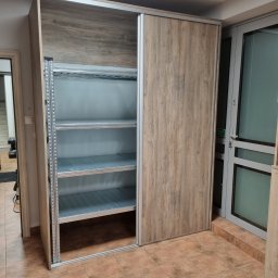 MEBLOTER Paweł Trytek - Szafa przesuwna z drewnopodobnymi drzwiami, częściowo otwarta, ukazująca metalowe półki. Widoczna struktura drewna na frontach i metalowe elementy konstrukcyjne.