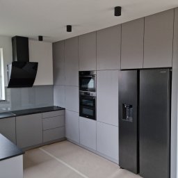 MEBLOTER Paweł Trytek - Nowoczesna kuchnia w odcieniach szarości z czarnymi akcentami: okap, piekarnik i lodówka side-by-side. Minimalistyczny design, gładkie fronty szafek, punktowe oświetlenie.