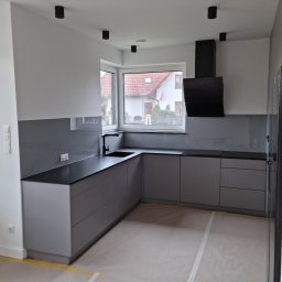 MEBLOTER Paweł Trytek - Nowoczesna, szara kuchnia z czarnym blatem i okapem, widok na dom przez okno. Podłoga zabezpieczona folią malarską, minimalistyczne oświetlenie sufitowe.