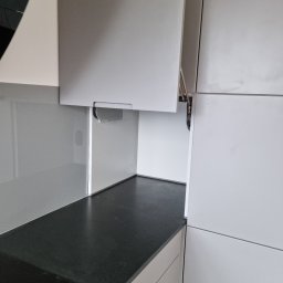 MEBLOTER Paweł Trytek - Nowoczesna kuchnia z szarymi szafkami i czarnym blatem. Szafka górna otwarta, ukazująca mechanizm podnoszenia. Minimalistyczny design, gładkie fronty.