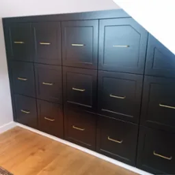 Czarna szafa wnękowa z szesnastoma drzwiczkami i złotymi uchwytami, dopasowana do skosu poddasza, widoczna drewniana podłoga i fragment dywanu.