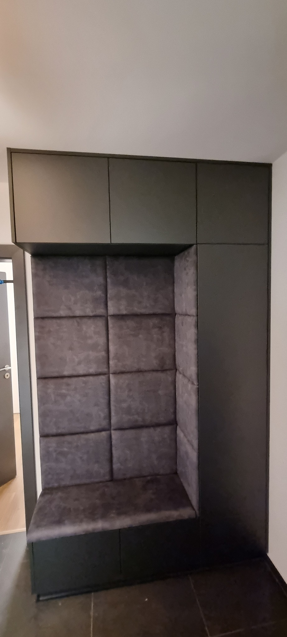 Tapicerowany narożnik wnękowy z szafkami górnymi w kolorze grafitowym. Miękkie panele ścienne w odcieniach szarości. Nowoczesny design, idealny do przedpokoju lub salonu.