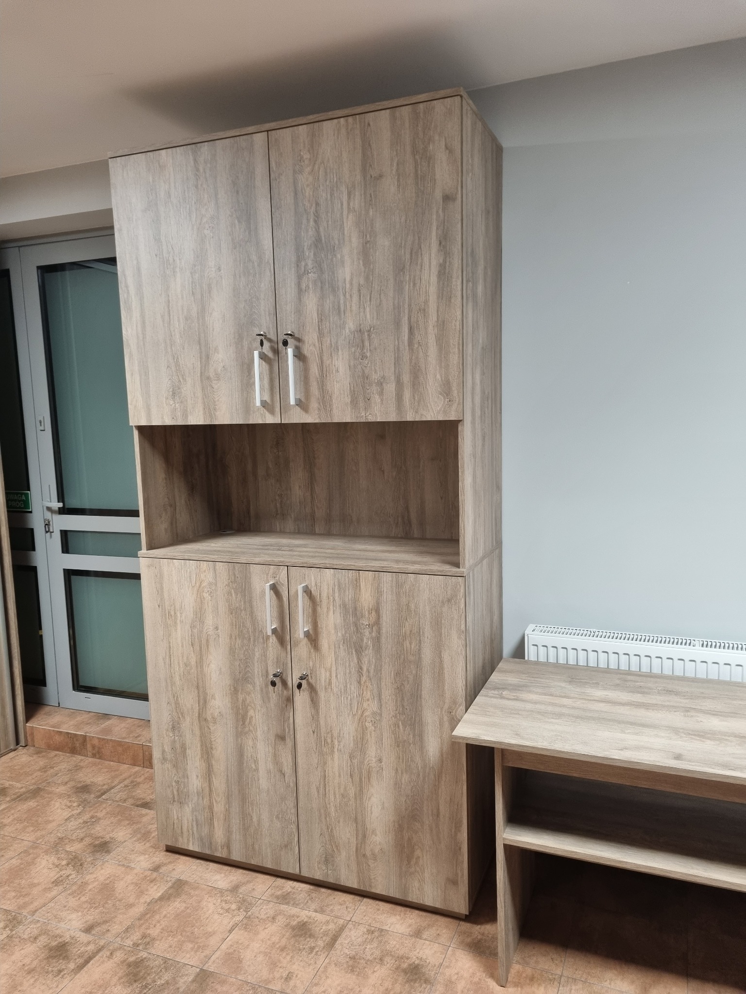 Wysoka szafa z półką i szafkami w odcieniach drewna, z metalowymi uchwytami, stoi na brązowej podłodze, obok stolik w podobnym stylu. Widoczne drzwi balkonowe.