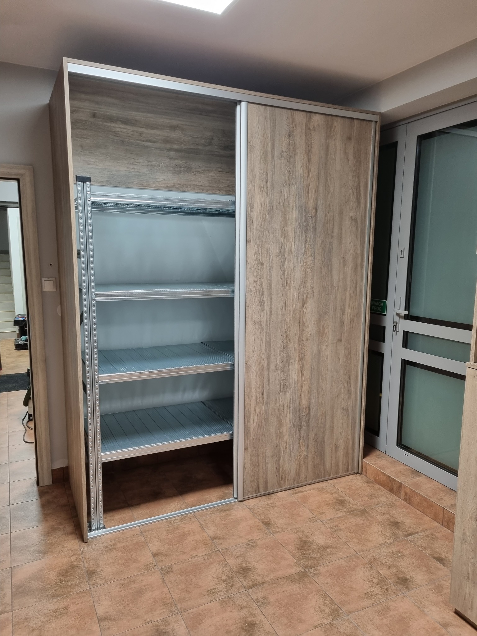 Szafa przesuwna z drewnopodobnymi drzwiami, częściowo otwarta, ukazująca metalowe półki. Widoczna struktura drewna na frontach i metalowe elementy konstrukcyjne.