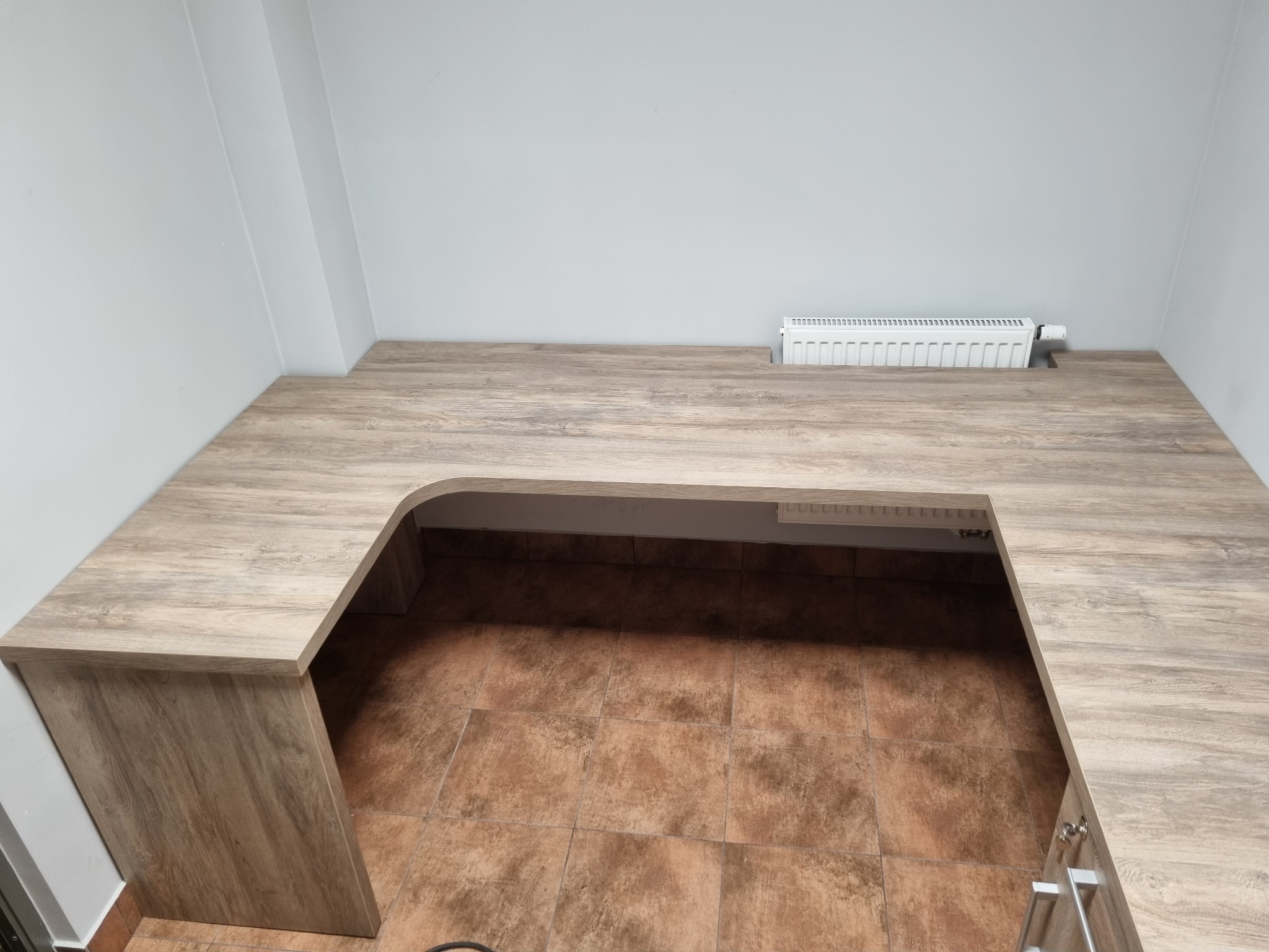 Blat drewniany w kształcie litery U, z wycięciem na grzejnik, zamontowany w rogu pomieszczenia z brązowymi płytkami podłogowymi. Widok z góry.